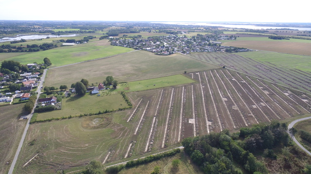 En mark med arkæologiske søgegrøfter. I baggrunden ses St. Rørbæk og Roskilde Fjord. Foto: Frederikssund Kommune.